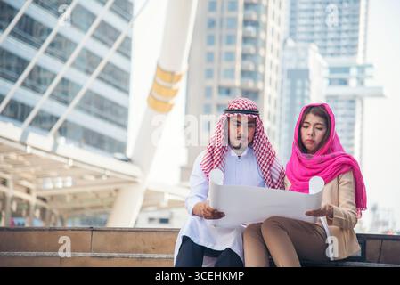 Arab civil construction engineer team consulting project hold blueprint pointing at construction site in muslim UAE city. Diversity teamwork manager p Stock Photo