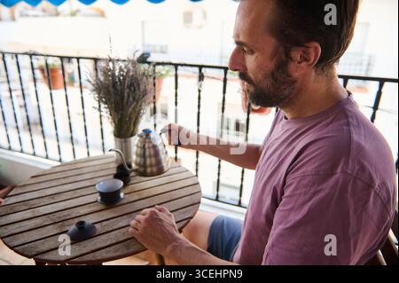 A man is seated outdoors on a warm balcony, engaged in preparing pour-over coffee. The warm sunlight, cozy wooden table setup, and a relaxing ambiance Stock Photo