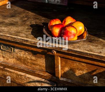 Several ripe nectarines are arranged on a dark ceramic plate, placed on a weathered wooden surface, bathed in warm sunlight, creating a still life com Stock Photo