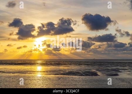 Sun setting behind clouds over calm sea at low tide, creating golden light beams and reflections Stock Photo
