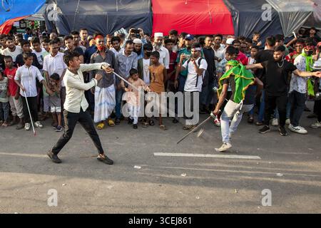 Chattogram, Bangladesh - 29 July 2023: View of two young men engage in a spirited mock sword fight amidst a captivated crowd, their movements a blur of white and green against the gray asphalt. Stock Photo