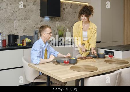 In a contemporary kitchen, a couple is sharing a moment as one partner serves a meal while the other is seated. The setting features stylish decor and Stock Photo