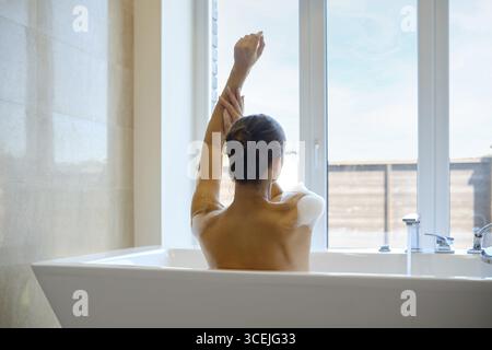 An unrecognizable woman enjoys a tranquil moment in a spacious bathtub while extending her arm. Sunlight streams in through large windows, enhancing t Stock Photo