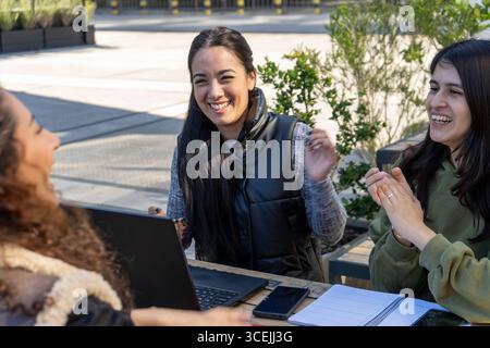Three happy university students are laughing and clapping while studying together at an outdoor table with laptops and notebooks Stock Photo
