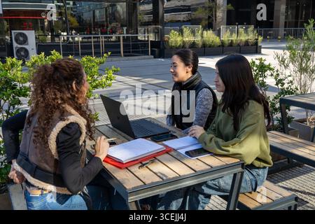 Three university students are working together on a project at an outdoor table, using a laptop and taking notes in their notebooks Stock Photo