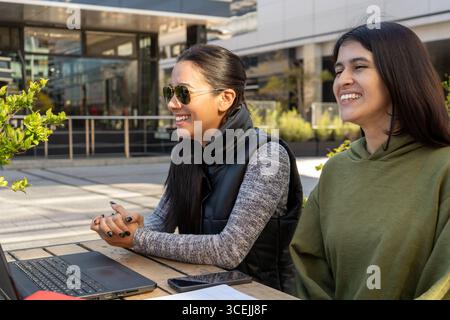 Two happy businesswomen are working remotely on a laptop while sitting at a table outside Stock Photo