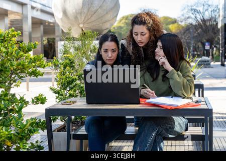 Three university students collaborating on a project outdoors, using a laptop and taking notes Stock Photo