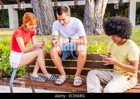 Young students using smartphones sitting on bench in park Stock Photo