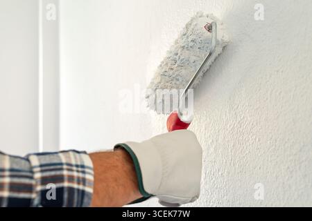 Close-up of hand wearing protective gloves. A painter paints a masonry wall with white paint using a roller. Construction industry. Safety at Work. Stock Photo