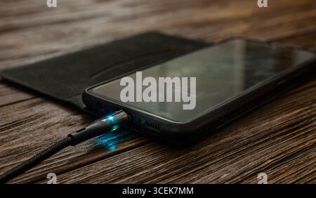A close-up shot of a smartphone connected to a glowing charging cable on a rustic wooden table, highlighting modern technology and connectivity. Stock Photo