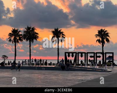 This photo captures a public square near the sea during sunset. Several people are walking, sitting, and playing. The large WHERE installation is in t Stock Photo