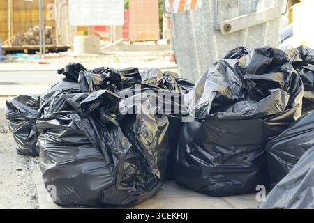 Black garbage bags piled on the street near dumpster awaiting collection. Symbol of urban waste, pollution, city cleanup, ecology and modern issues Stock Photo