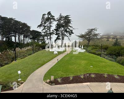 Elegant outdoor wedding and event setup with white tables, chairs, and umbrellas arranged on a lawn overlooking the Pacific Ocean on a foggy day Stock Photo