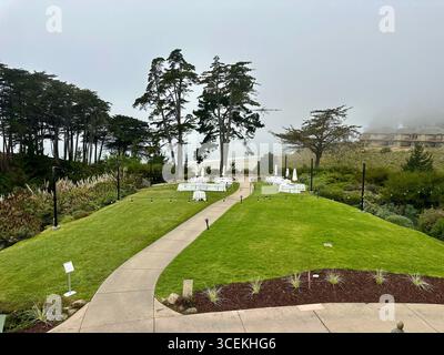 Elegant outdoor wedding and event setup with white tables, chairs, and umbrellas arranged on a lawn overlooking the Pacific Ocean on a foggy day Stock Photo