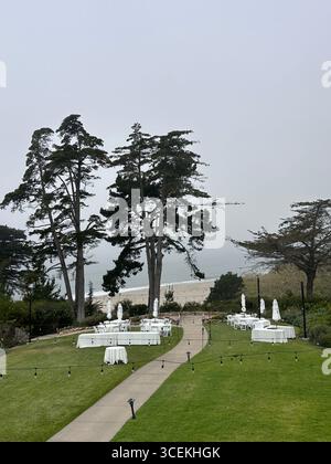 Elegant outdoor wedding and event setup with white tables, chairs, and umbrellas arranged on a lawn overlooking the Pacific Ocean on a foggy day Stock Photo