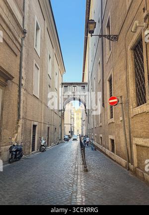 A long, narrow cobblestone street, lined with tall brick buildings and a unique overhead archway in Rome, Italy. Parked scooters and a few people walk Stock Photo