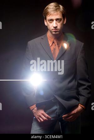 A man in a suit and tie stands with his hands in his pockets, looking directly at the camera. A bright light shines in front of him. Stock Photo