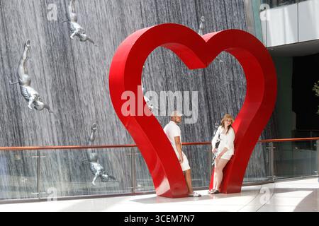 Couple posing in front of a large red heart sculpture near a waterfall with statues of divers at dubai mall shopping centre UAE Stock Photo