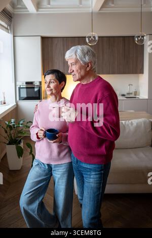 Senior couple watching street through window, talking and drinking tea. Community gossip exchange Stock Photo