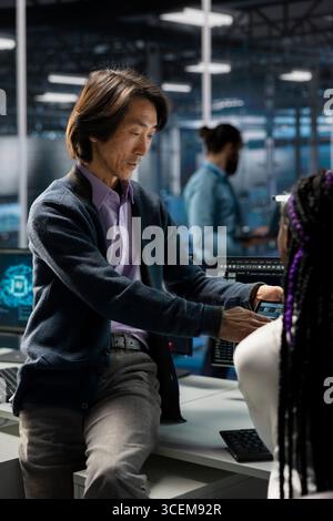Manager and intern in data center troubleshoot server connectivity issues using AI. Senior programmer and young colleague in server room audit systems for vulnerabilities using artificial intelligence Stock Photo