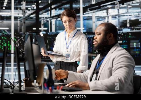 Data center manager supervising engineer inspecting gear, doing maintenance tasks. Server farm team leader supervising employee conducting system checks to ensure operational stability across rigs Stock Photo