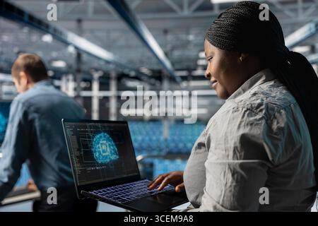 Smiling IT expert in server room using laptop to improve AI system scalability for increased workloads. Happy data center programmer using notebook to adjust artificial intelligence model parameters Stock Photo