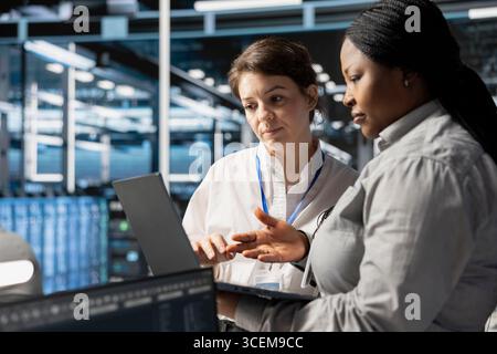 Team of system administrators in data center using laptop to update storage arrays software, configuring network virtualization. Server hub IT specialists using notebook to verify settings Stock Photo