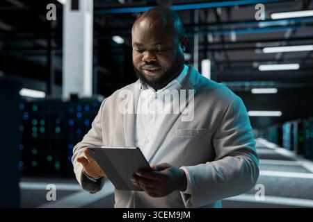 Engineer in server farm coordinating backups protocols, checking recovery plan. Data center IT specialist performing maintenance with tablet, making sure rigs are functioning optimally Stock Photo