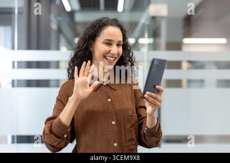 A cheerful woman waves while using her smartphone for a video conversation, seated in a contemporary office environment with glass panels in the background, highlighting connection Stock Photo