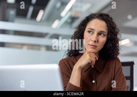 A woman in a brown outfit sitting thoughtfully at a desk in a contemporary office, surrounded by a clean and modern environment, with paperwork and a laptop symbolizing work or problem-solving. Stock Photo