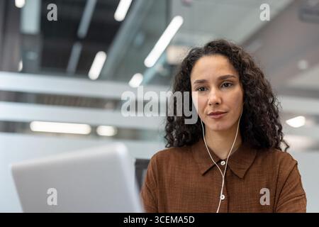 A focused professional sits at a desk, wearing headphones while engaged in focused work on her laptop. The setting is a modern office with stylish details and bright light. Stock Photo