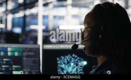 Data center employee uses audio headset and visual programming dashboard to troubleshoot client reported issues. Server farm technician looks at node tree interface, talking in microphone Stock Photo