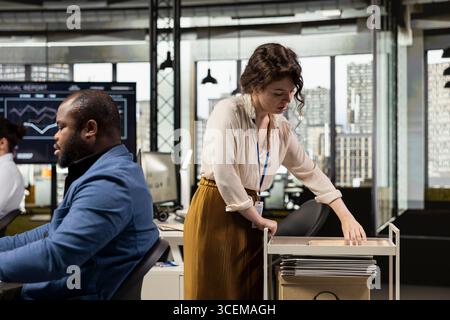 Workplace support employee moves a delivery cart stacked with folders and contracts, helping african american administrator bringing stack of archived documents for accounting tasks. Stock Photo