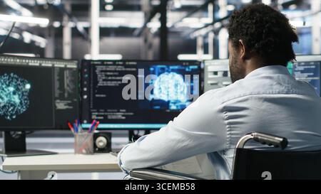 Server room programmer with paraplegia interprets AI generated data analytics graphs. Man in wheelchair using artificial intelligence program showing predictive system metrics in data center, camera B Stock Photo