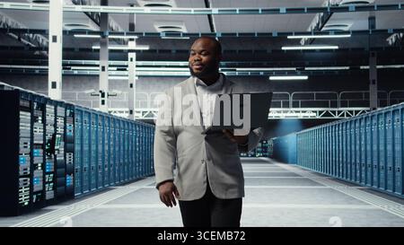 Smiling server room engineer performing maintenance on server racks using software on laptop. Happy data center IT professional upgrading blade server components using notebook, camera A Stock Photo