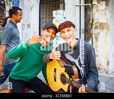 Two young asian men smiling giving thumbs up, one holding a guitar, in a  street buskers musicians George town Penang Malaysia Stock Photo