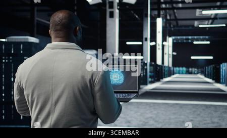 IT expert walking in server hub using laptop to enhance AI model accuracy. Data center technician using notebook to schedule routine maintenance for AI hardware components, camera A Stock Photo