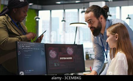 Young programmer in office requests help from IT experts detecting and blocking hacker intrusion. Intern assisted by staff members coordinating incident response for network attack, camera A Stock Photo