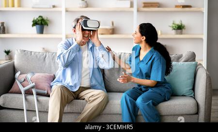 Elderly man using VR headset with young medical worker, exploring augmented reality at retirement home Stock Photo