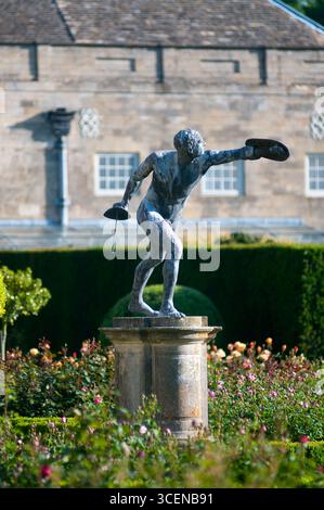 A dancer statue captures a dynamic pose in a vibrant rose garden, framed by neatly trimmed hedges and elegant historic buildings under blue skies. Stock Photo