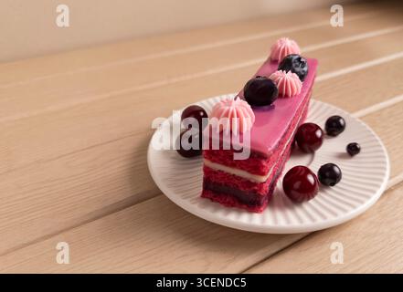 A delicious slice of berry layer cake topped with pink cream, cherries and blueberries, served on a white plate on a wooden table. Perfect for dessert Stock Photo
