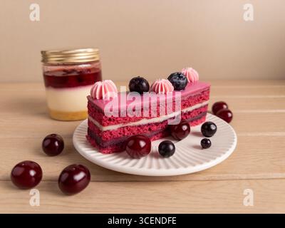 A slice of layered berry cake with pink frosting and fresh berries, artfully presented on a white plate. In the background, a jar of layered dessert a Stock Photo