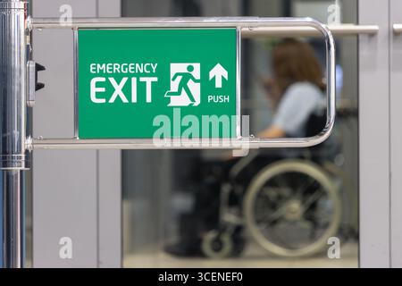 Green emergency exit sign on metal gate indoors with disabled person in wheelchair on blurred background Stock Photo