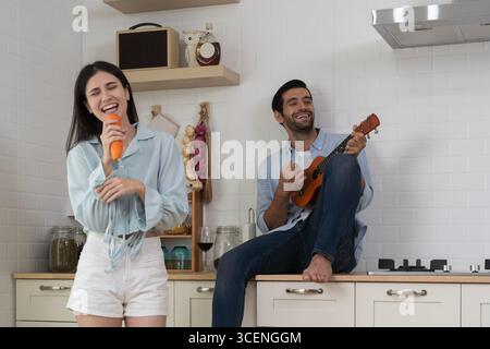 Young couple enjoying fun moment at home, woman singing with carrot as microphone, man playing ukulele, cheerful lifestyle, happiness, love, relations Stock Photo
