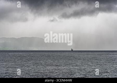 A ferry moves steadily across the choppy sea as dark clouds gather, hinting at an approaching storm in the distance. Stock Photo