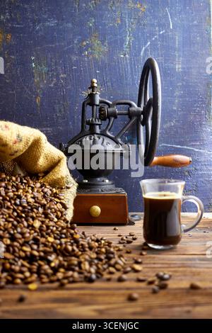 Rustic coffee still life with burlap sack spilling roasted beans, vintage grinder, and glass cup of fresh coffee on a wooden table, styled in farmhous Stock Photo