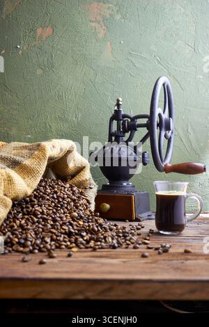 Rustic coffee scene with vintage grinder, burlap sack spilling roasted beans, and glass cup of black coffee on a wooden table, styled in farmhouse kit Stock Photo