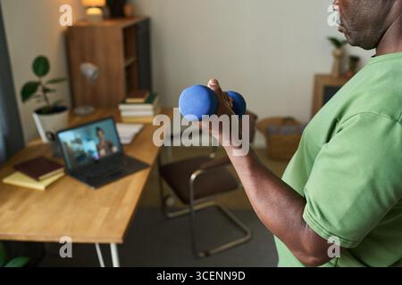 Senior Black man exercising with dumbbell while following online fitness class on laptop in home office, focusing on strength training and maintaining healthy lifestyle Stock Photo