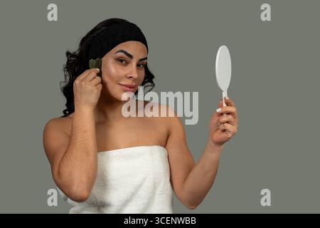 Woman using a gua sha stone and holding a mirror during her skincare routine, promoting beauty and self-care Stock Photo