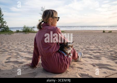 Woman relaxing on sandy beach with cat on leash at morning walk rear view. Feline curious about sand Stock Photo